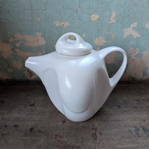 May include: A white ceramic teapot with a unique, curved design. The teapot has a matching lid with a small handle. The handle on the teapot is a curved loop. The teapot is sitting on a distressed, dark surface.