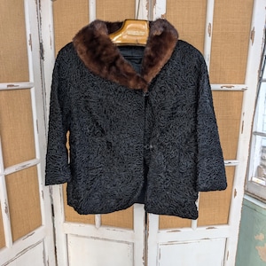 May include: Black vintage jacket with a textured pattern and a brown fur collar. The jacket has three-quarter sleeves and is buttoned in the front. The jacket is hanging on a wooden hanger.
