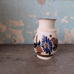 May include: A small, white ceramic vase with a blue and brown floral design. The vase has a rounded body and a narrow neck.