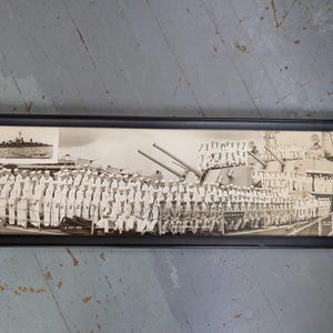 May include: Black and white framed photograph of a naval ship and crew. The image shows a large group of sailors in uniform standing on the deck of a ship. A smaller image of a ship is in the upper left corner. The frame is black.