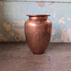May include: A polished copper vase with a rounded body, a narrow neck, and a flared rim. The surface of the metal shows subtle variations in texture and color, reflecting light across its curved form. The vase has a wide base and a smooth finish.
