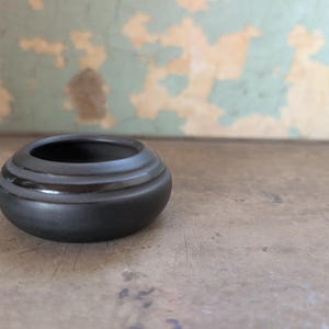May include: A small, round, matte black ceramic bowl with a low profile and a wide, open top. The bowl features a smooth, curved exterior surface with a subtle horizontal ridge detail encircling the upper portion near the rim.