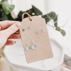 May include: A beige leather earring display card with silver earrings in various shapes, including stars, geometric patterns, and circles.