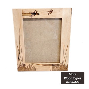 May include: A wooden picture frame with a natural wood finish. The frame features a woodburned design of ducks flying over a marsh with cattails. The text "More Wood Types Available" is printed below the frame.
