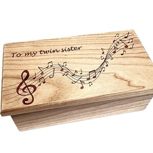 May include: A wooden box with a light brown finish. The lid is engraved with the words "To my twin sister" and a design of musical notes and a treble clef.