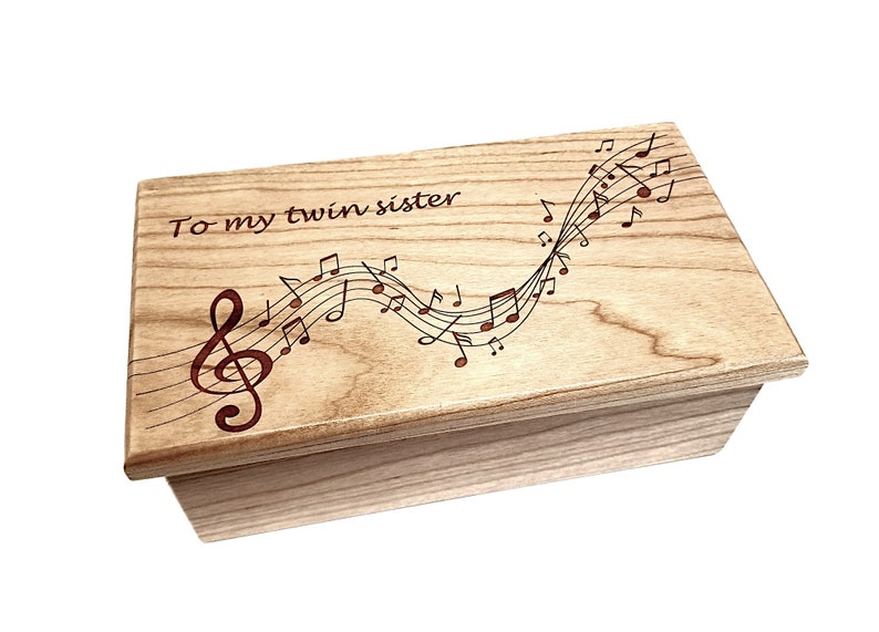 May include: A wooden box with a light brown finish. The lid is engraved with the words "To my twin sister" and a design of musical notes and a treble clef.