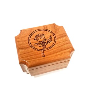 Puede incluir: Pequeña caja de madera cuadrada con tapa con bisagras. La caja es de madera marrón claro y presenta un diseño grabado con láser de una rosa dentro de un marco circular en la parte superior. Las esquinas de la caja son redondeadas.