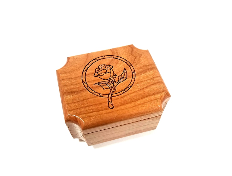May include: A small, square wooden box with a hinged lid. The box is made of light brown wood and features a laser-etched design of a rose within a circular frame on the top. The corners of the box are rounded.