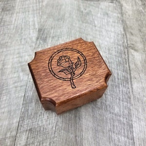 May include: A small, wooden box with a dark brown finish. The lid features a laser-etched design of a rose within a circular frame. The box has a unique, scalloped edge design.