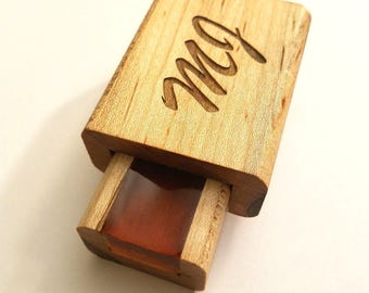 May include: A wooden box with a sliding drawer, featuring the engraved letters "mQ" on the top. The drawer is partially open, revealing a reddish-brown interior. The box is made of light-colored wood.