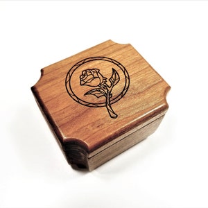 May include: A small, square wooden box with a hinged lid. The box is made of light brown wood and features a laser-etched design of a rose within a circular frame on the top. The box has a decorative, scalloped edge.