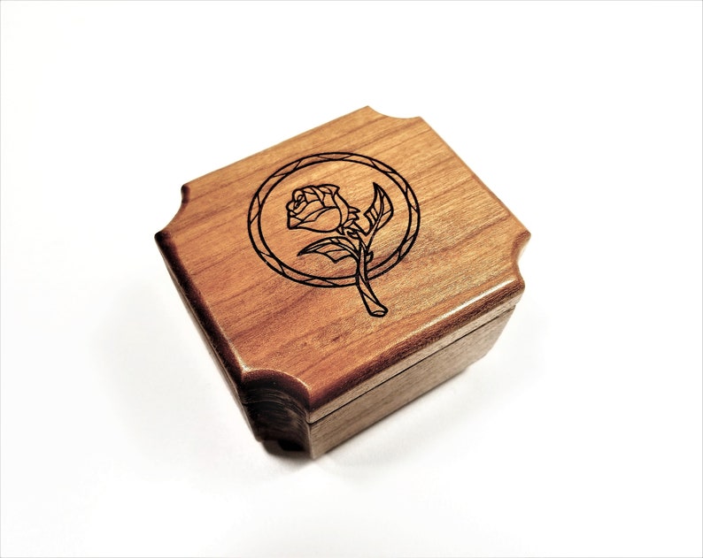 May include: A small, square wooden box with a hinged lid. The box is made of light brown wood and features a laser-etched design of a rose within a circular frame on the top. The box has a decorative, scalloped edge.