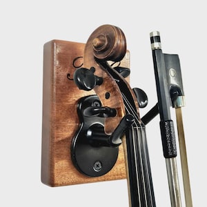 May include: A wooden violin wall mount with a black metal bracket holding a violin. The violin's strings and bow are visible. The mount is made of dark wood and is mounted on a white wall.