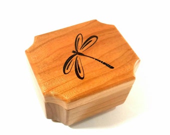 Puede incluir: Una pequeña caja de madera con un diseño de libélula tallado en la tapa. La caja está hecha de madera marrón claro y tiene una forma ligeramente redondeada.