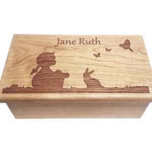 May include: A wooden box with a personalized inscription "Jane Ruth" and an engraved design of a child sitting with a rabbit and butterflies. The box is made of light wood with a natural finish.