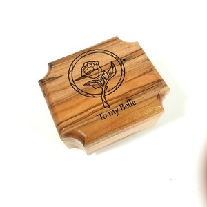 May include: A wooden box with a rose illustration and the text "To my Belle". The box has a light brown color with a natural wood grain pattern. The rose is inside a circle. The box has a unique, curved edge design.