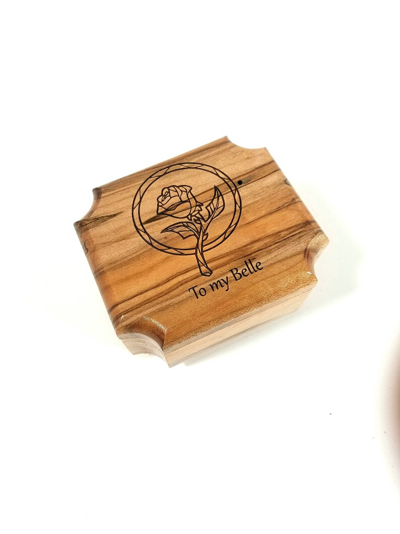 May include: A wooden box with a rose illustration and the text "To my Belle". The box has a light brown color with a natural wood grain pattern. The rose is inside a circle. The box has a unique, curved edge design.