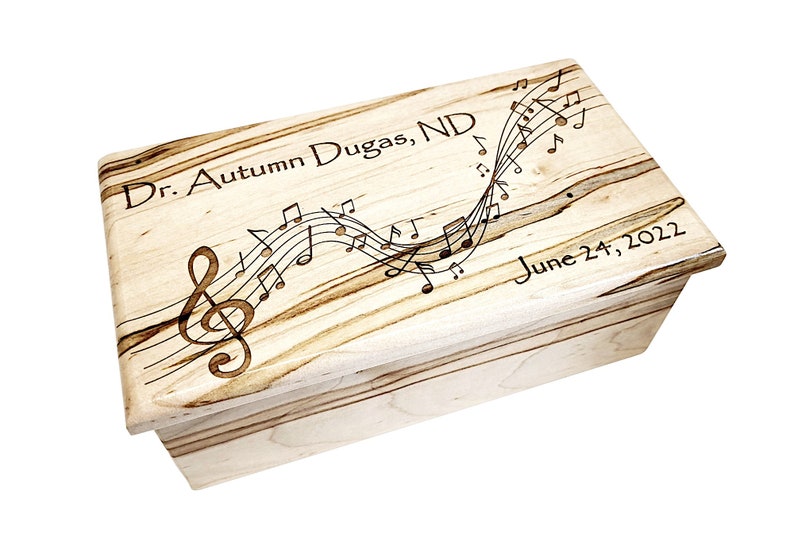 May include: A wooden box with a light brown wood grain finish. The top of the box is engraved with musical notes and a treble clef. The text "Dr. Autumn Dugas, ND" and "June 24, 2022" is also engraved on the box.