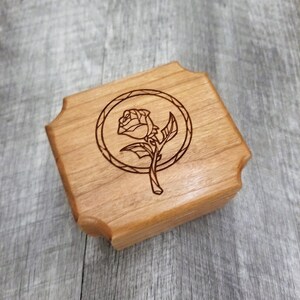 May include: A small, square wooden box with a carved rose design inside a circle. The box is made of light-colored wood and has decorative, curved edges. The rose is depicted with detailed lines.