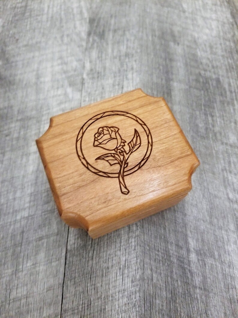 May include: A small, square wooden box with a carved rose design inside a circle. The box is made of light-colored wood and has decorative, curved edges. The rose is depicted with detailed lines.
