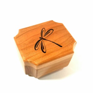 May include: A small wooden box with a dragonfly design carved into the lid. The box is made of light brown wood and has a slightly rounded shape.