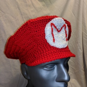 May include: Red crocheted hat with a white circle and a red letter M on top. The hat is on a black mannequin head.