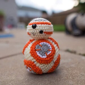 May include: A crocheted toy resembling the character BB-8 from the Star Wars franchise. The toy is white with orange and gray accents. The toy has a round body and a smaller, round head.