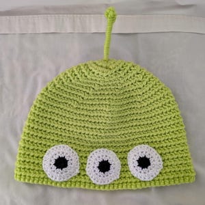 May include: A lime green crocheted hat with three white and black eye appliqués. The hat has a small antenna on top. The hat is likely for a child.