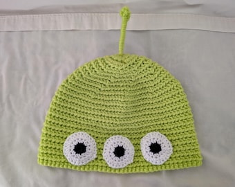 May include: A lime green crocheted hat with three white and black eye appliqués. The hat has a small antenna on top. The hat is likely for a child.