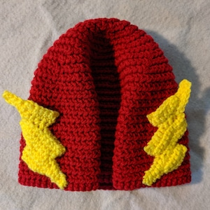 May include: A handmade red crochet hat with two yellow lightning bolt appliqués on the sides. The hat is a beanie style, likely for a child or baby. The yarn appears to be a medium weight.