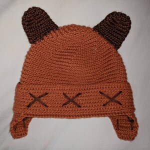 May include: A brown crocheted hat with brown ear flaps and brown X-shaped stitching on the brim.