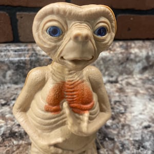 May include: A tan-colored E.T. figurine with large blue eyes and a distinctive chest marking. The figure is holding its fingers up, a classic pose from the movie. A small, circular accessory is attached.