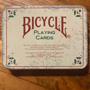 Puede incluir: Una lata rectangular de naipes Bicycle. La lata tiene un fondo color crema con elementos decorativos rojos y verdes. Las palabras "BICYCLE PLAYING CARDS" se muestran de forma destacada en una fuente vintage.