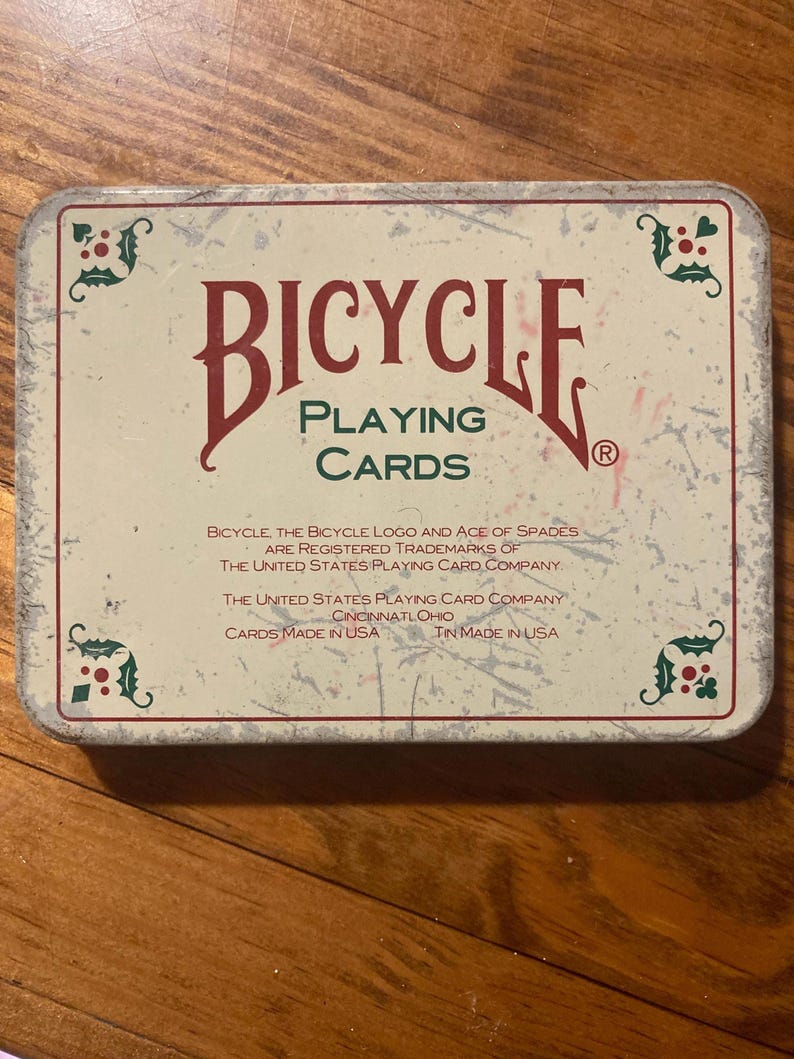 Puede incluir: Una lata rectangular de naipes Bicycle. La lata tiene un fondo color crema con elementos decorativos rojos y verdes. Las palabras "BICYCLE PLAYING CARDS" se muestran de forma destacada en una fuente vintage.