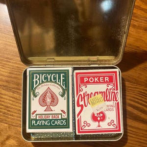Puede incluir: Una lata dorada abierta que contiene dos barajas de cartas. Una baraja es verde y blanca, etiquetada como "Bicycle Holiday Back Playing Cards". La otra es roja y blanca, etiquetada como "Poker Streamline Playing Cards".