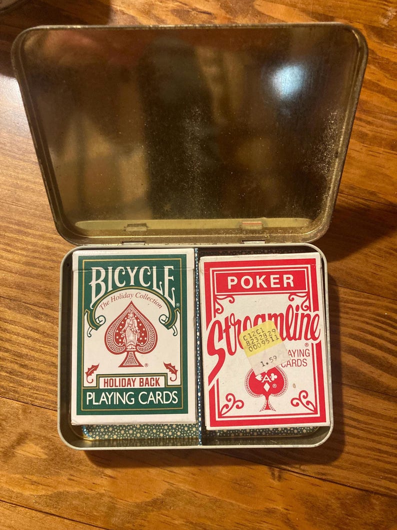 Puede incluir: Una lata dorada abierta que contiene dos barajas de cartas. Una baraja es verde y blanca, etiquetada como "Bicycle Holiday Back Playing Cards". La otra es roja y blanca, etiquetada como "Poker Streamline Playing Cards".