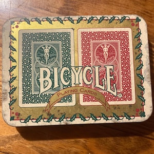 Puede incluir: Una lata rectangular de naipes Bicycle con un dise&ntilde;o de cartas verdes y rojas. La lata presenta la palabra "Bicycle" en letras blancas grandes, con una pancarta que dice "Playing Cards". La lata est&aacute; decorada con un borde dorado y hojas de acebo.