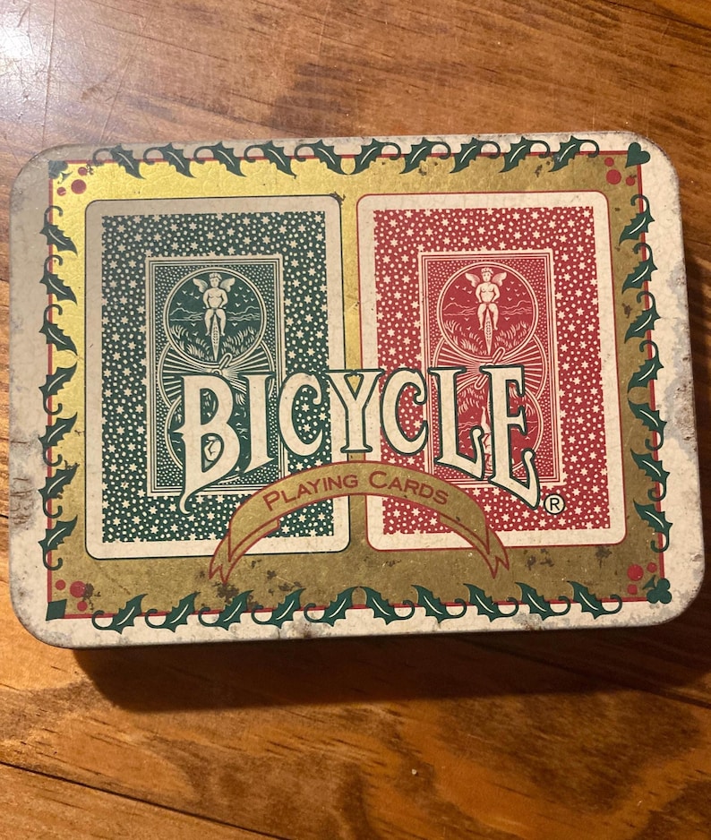 Puede incluir: Una lata rectangular de naipes Bicycle con un dise&ntilde;o de cartas verdes y rojas. La lata presenta la palabra "Bicycle" en letras blancas grandes, con una pancarta que dice "Playing Cards". La lata est&aacute; decorada con un borde dorado y hojas de acebo.