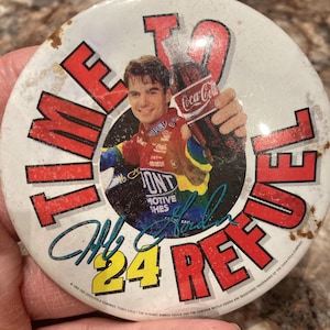 May include: A round, vintage Coca-Cola button featuring a smiling man holding a Coca-Cola bottle. The button has the text "TIME 24 REF" in red, with the number 24 in yellow. The man's signature is in blue. The button has a distressed, aged appearance.