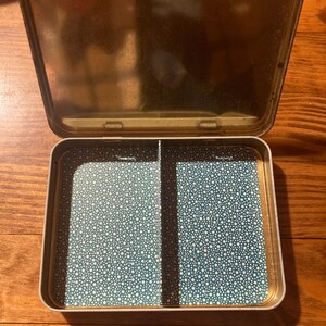 Puede incluir: Una caja met&aacute;lica rectangular abierta con tapa abisagrada. El interior presenta un patr&oacute;n de estrellas turquesas y blancas, dividido en dos compartimentos. La tapa y el exterior tienen un acabado dorado.