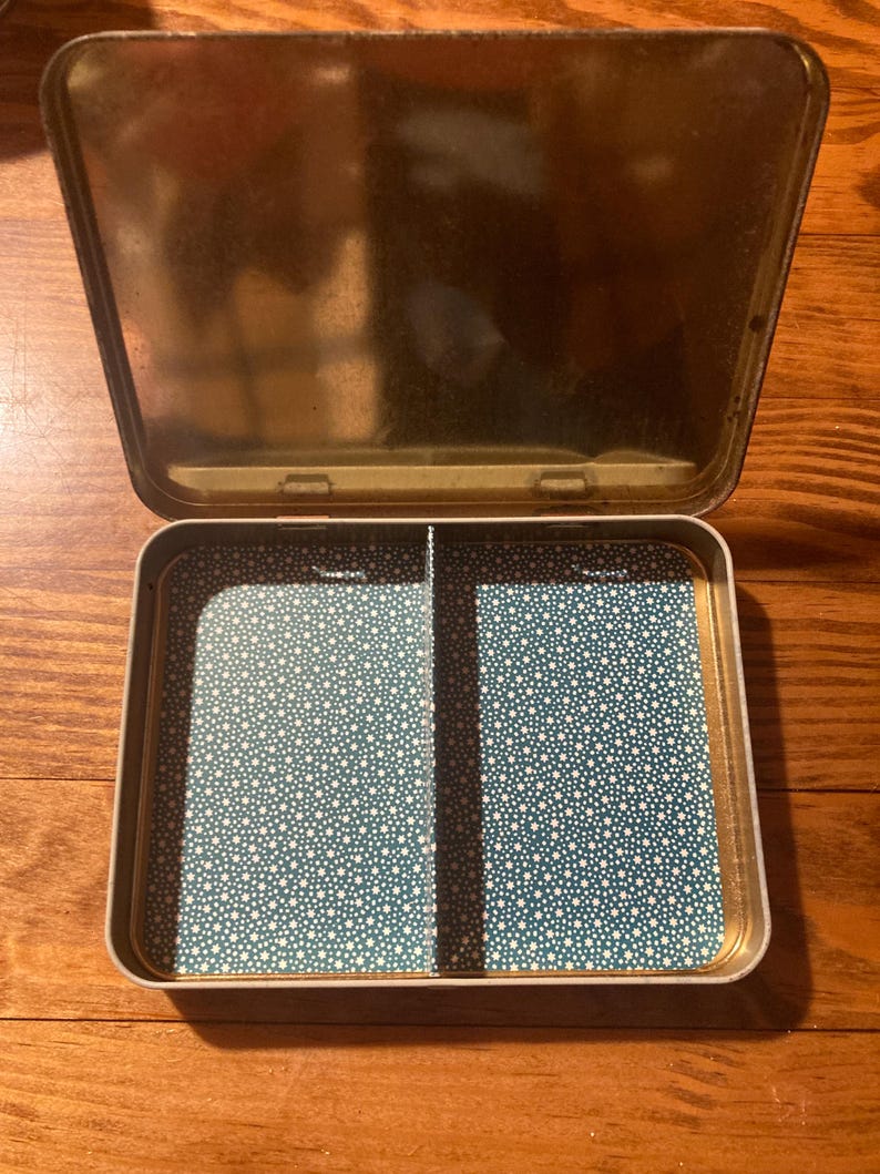 Puede incluir: Una caja met&aacute;lica rectangular abierta con tapa abisagrada. El interior presenta un patr&oacute;n de estrellas turquesas y blancas, dividido en dos compartimentos. La tapa y el exterior tienen un acabado dorado.