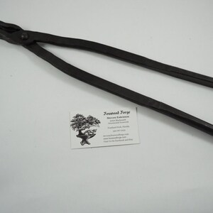 May include: Black metal blacksmith tongs with a rounded handle. The tongs are approximately 20 inches long.