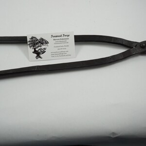 May include: Black metal blacksmith tongs with a rounded handle and a pointed jaw. The tongs are on a white surface with a business card for "Ironwood Forge" in the background.