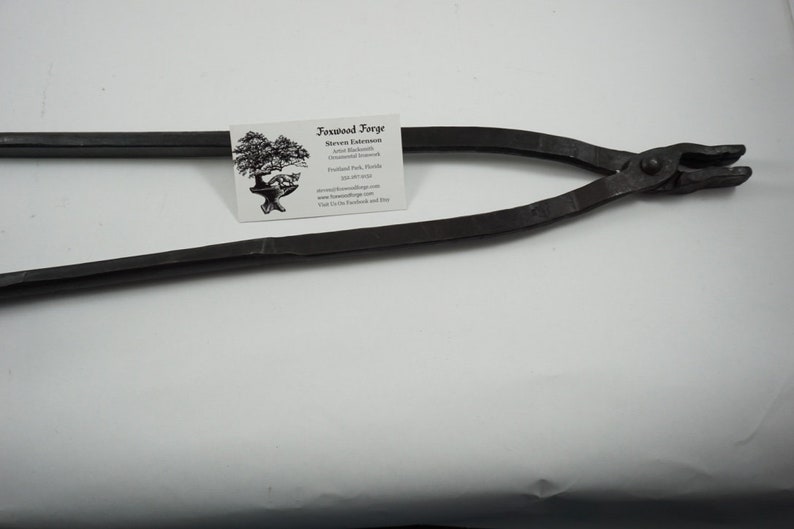 May include: Black metal blacksmith tongs with a rounded handle and a pointed jaw. The tongs are on a white surface with a business card for "Ironwood Forge" in the background.