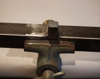 May include: A metal vise with a rectangular metal bar clamped in its jaws. The vise is painted blue-green and the metal bar is a dark gray color. The image is a close-up shot.