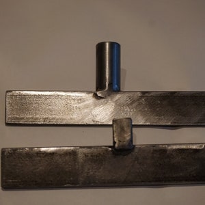 Op de afbeelding: Twee metalen platen met een metalen staaf die ze verbindt. De platen zijn rechthoekig en hebben een ruw, getextureerd oppervlak.