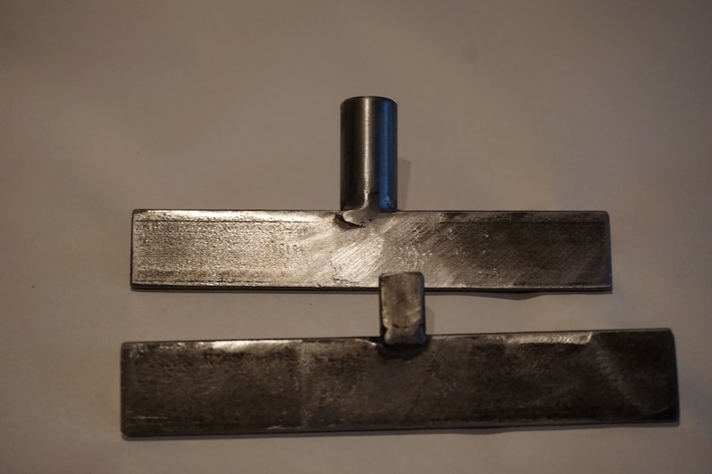 Op de afbeelding: Twee metalen platen met een metalen staaf die ze verbindt. De platen zijn rechthoekig en hebben een ruw, getextureerd oppervlak.