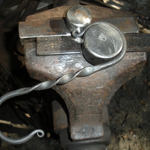 May include: A metal hook with a twisted design is clamped in a vise. The hook is made of dark metal and has a rounded end. The vise is made of metal and has a rough, textured surface.