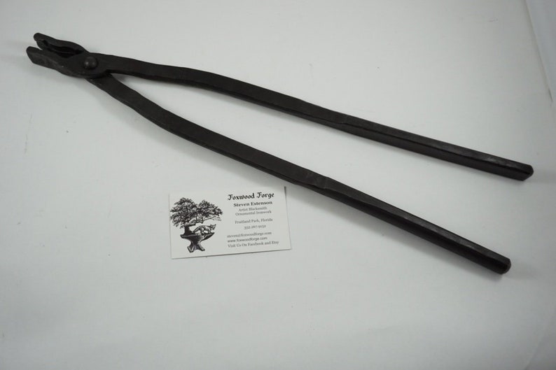 May include: Black metal blacksmith tongs with a rounded handle. The tongs are approximately 20 inches long.