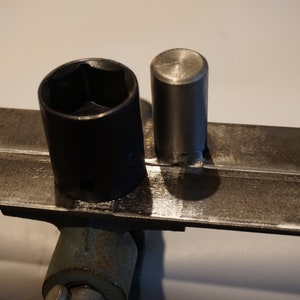 Op de afbeelding: Een close-up van twee metalen gereedschappen. Het gereedschap aan de linkerkant is een zwarte steeksleutel. Het gereedschap aan de rechterkant is een zilveren metalen staaf. Beide gereedschappen liggen op een metalen oppervlak.